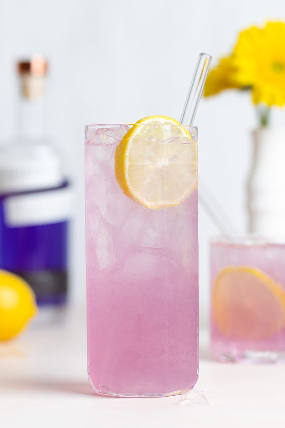 A light pink cocktail in a tall glass with ice garnished with a slice of lemon and a glass straw.