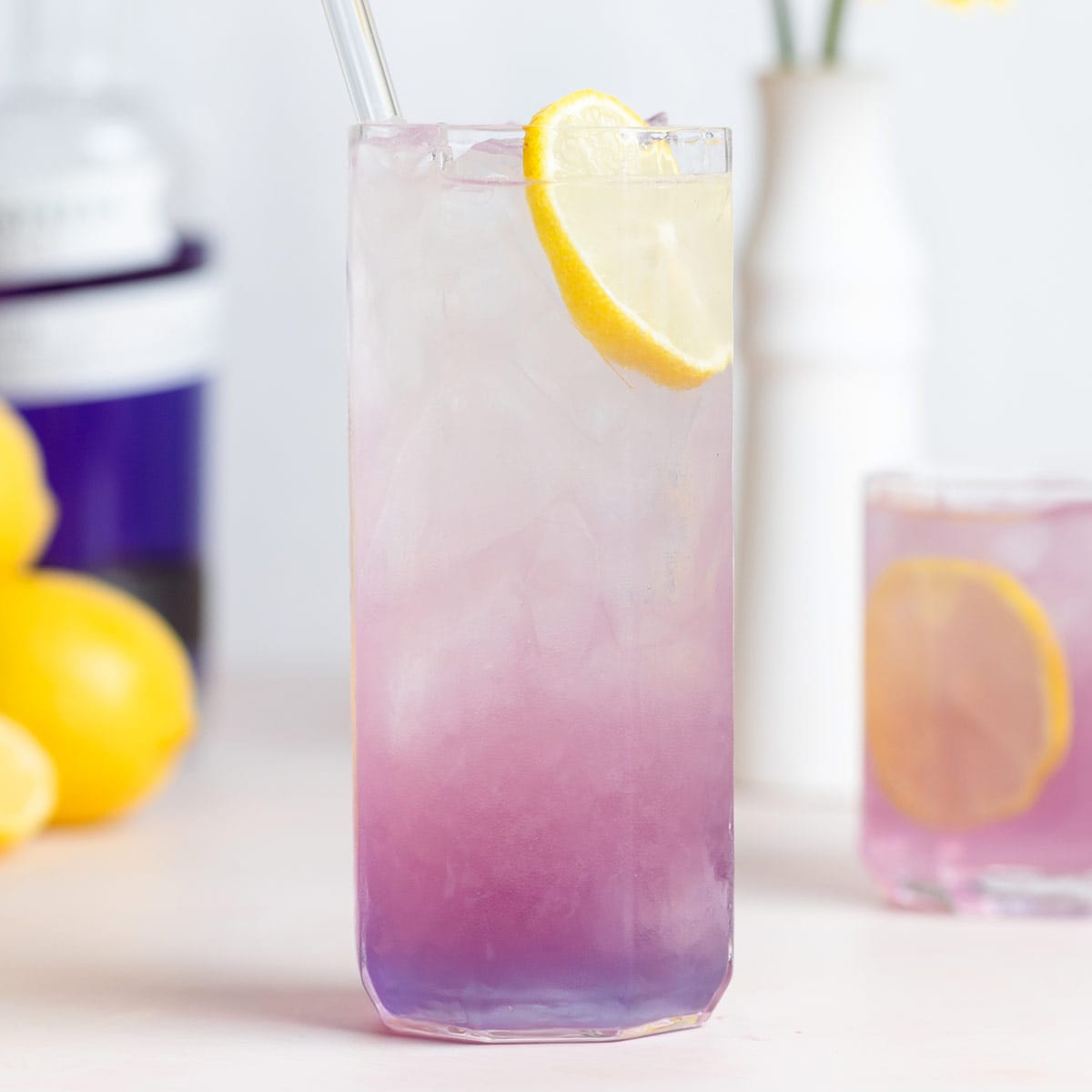 A light pink purple cocktail in a tall glass with an ombre effect garnished with a slice of lemon and a glass straw.
