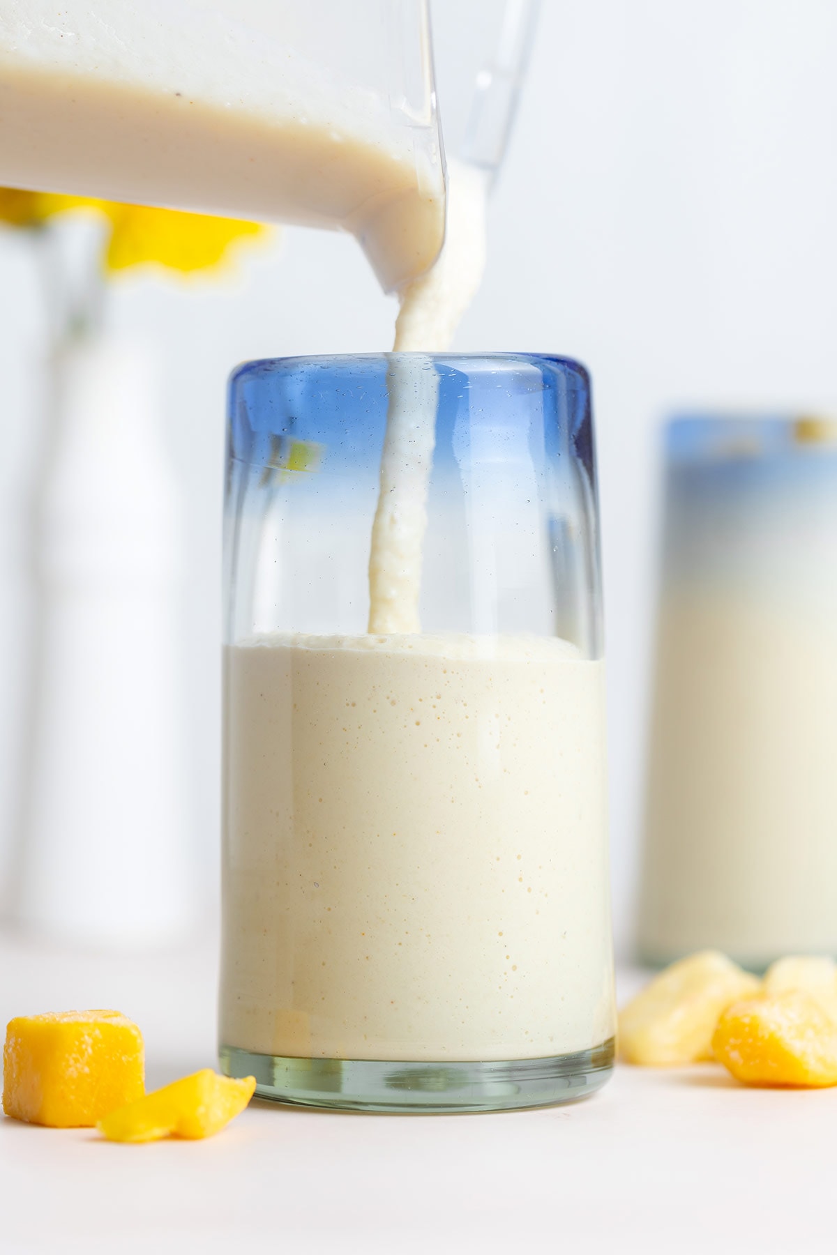 A light yellow smoothie being poured into a tall glass with a blue rim.