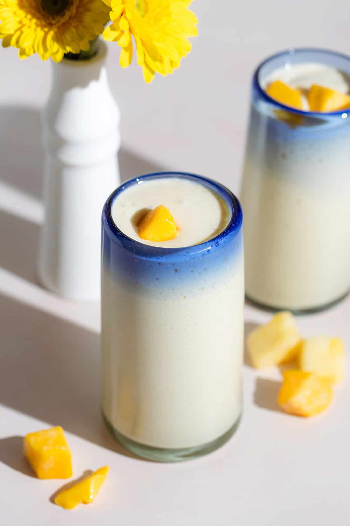 Light yellow smoothie in two tall glasses with a blue rim garnished with small pieces of mango.