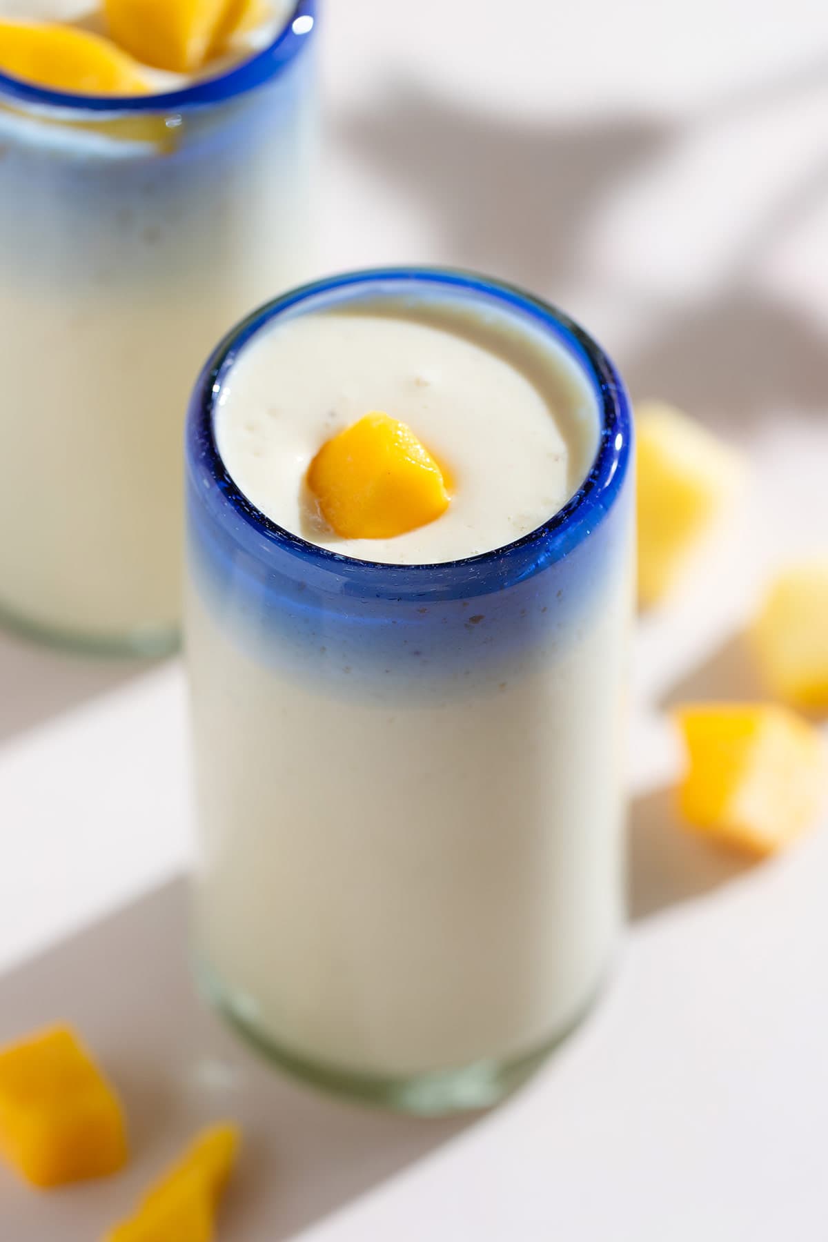 Light yellow smoothie in two tall glasses with a blue rim garnished with small pieces of mango.