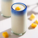 Light yellow smoothie in a tall glass with a blue rim garnished with small pieces of mango.