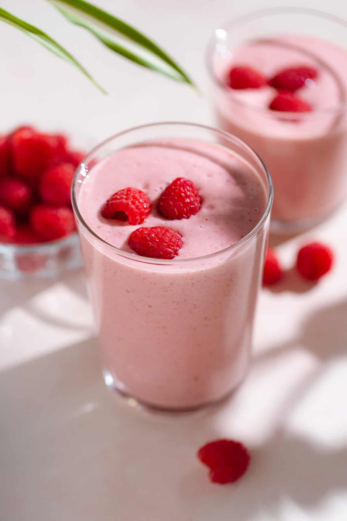 Creamy pink smoothie in a tall glass garnished with fresh raspberries on top.