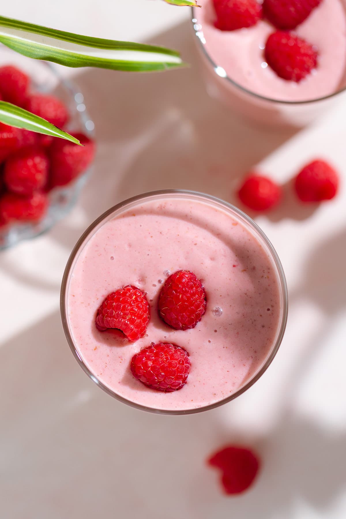 Creamy pink smoothie in a tall glass garnished with fresh raspberries on top.