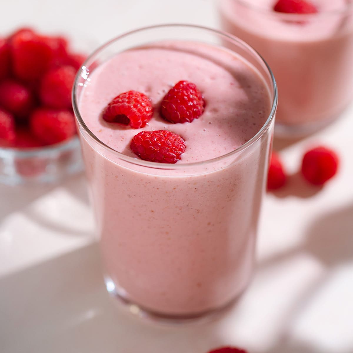Creamy pink smoothie in a tall glass garnished with fresh raspberries on top.