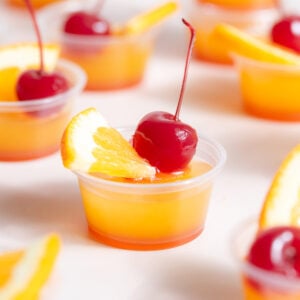 Small 2 oz plastic cups with tequila sunrise jello shots garnished with cocktail cherries and small orange slices.