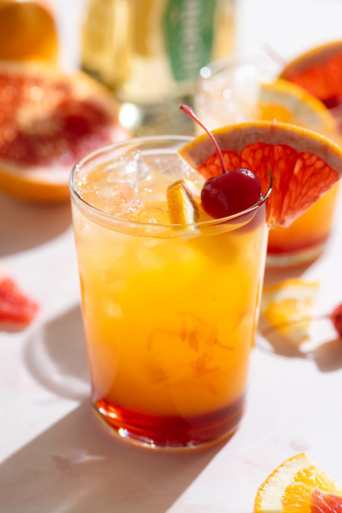 A tequila sunset cocktail in a tall glass with orange juice and red grenadine on the bottom of the glass for an ombre effect garnished with a cocktail cherry and a slice of grapefruit.