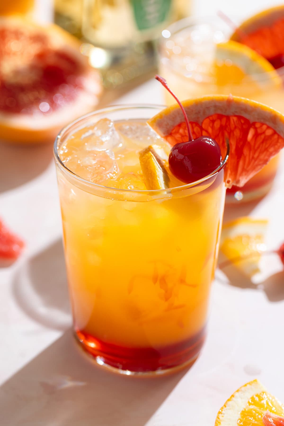 A tequila sunset cocktail in a tall glass with orange juice and red grenadine on the bottom of the glass for an ombre effect garnished with a cocktail cherry and a slice of grapefruit.
