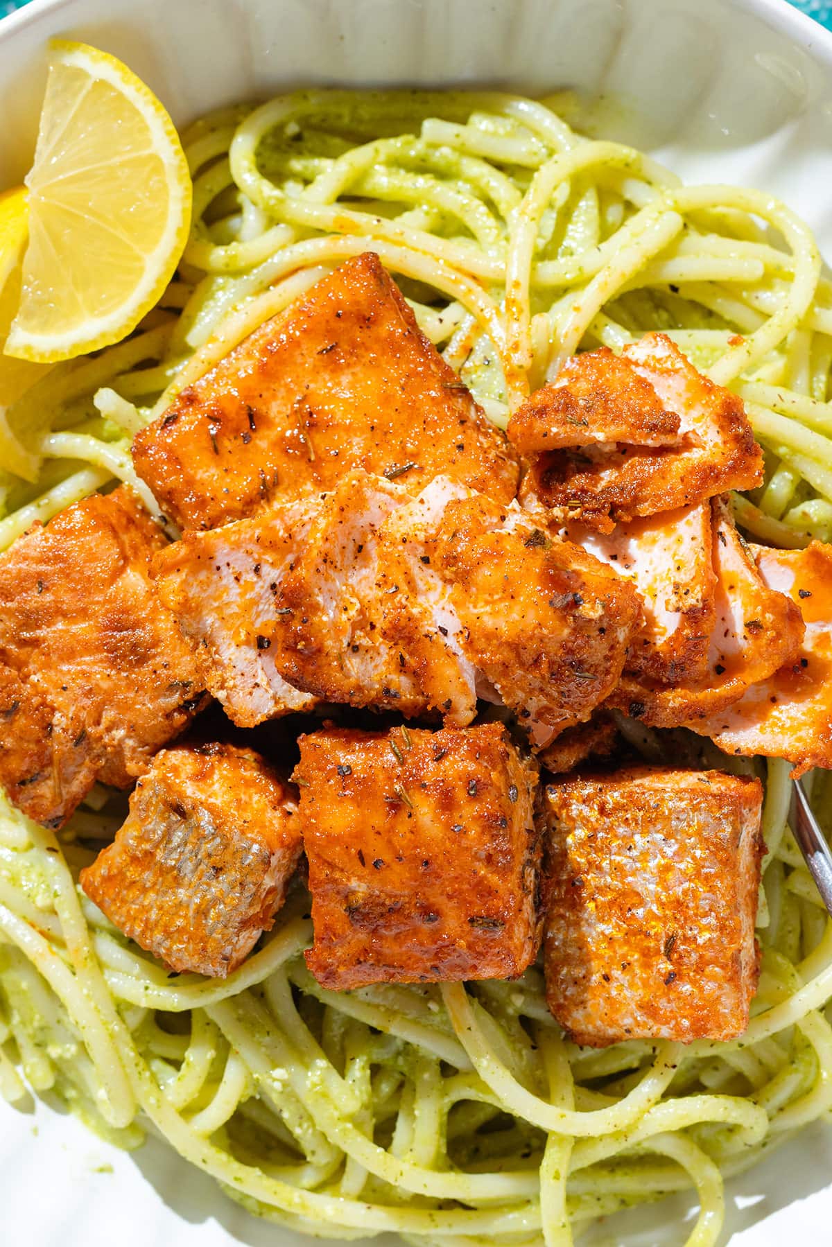 Pesto spaghetti in a white bowl topped with roasted diced salmon and lemon wedges on the side.