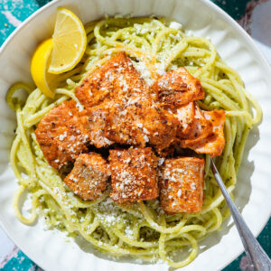 Pesto spaghetti in a white bowl topped with roasted salmon and pecorino cheese with lemon wedges on the side.