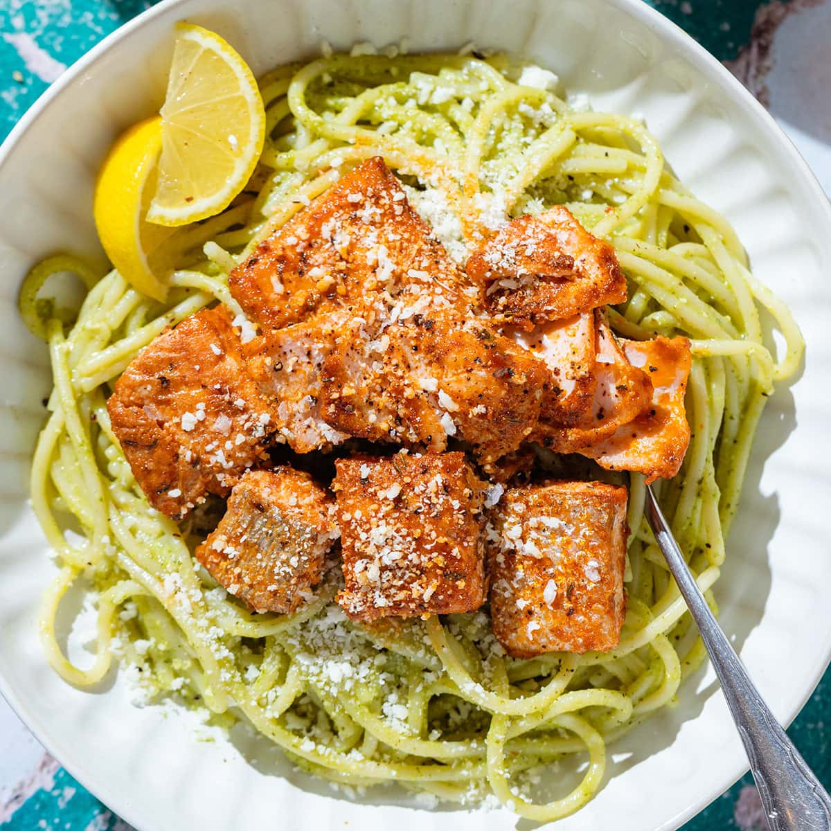 Pesto spaghetti in a white bowl topped with roasted salmon and pecorino cheese with lemon wedges on the side.