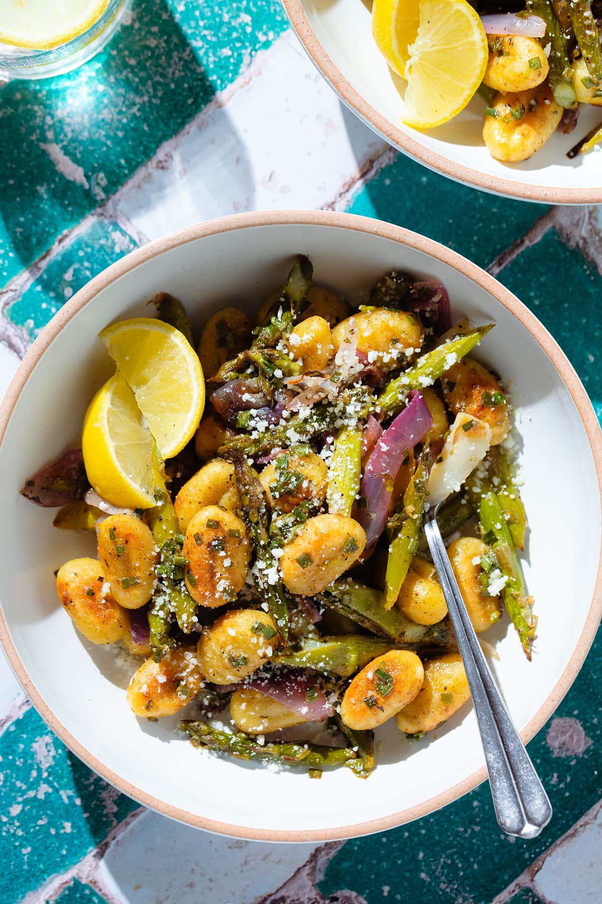 Crispy golden gnocchi with roasted asparagus, red onion, and pecorino in a white bowl with lemon wedges on the side.