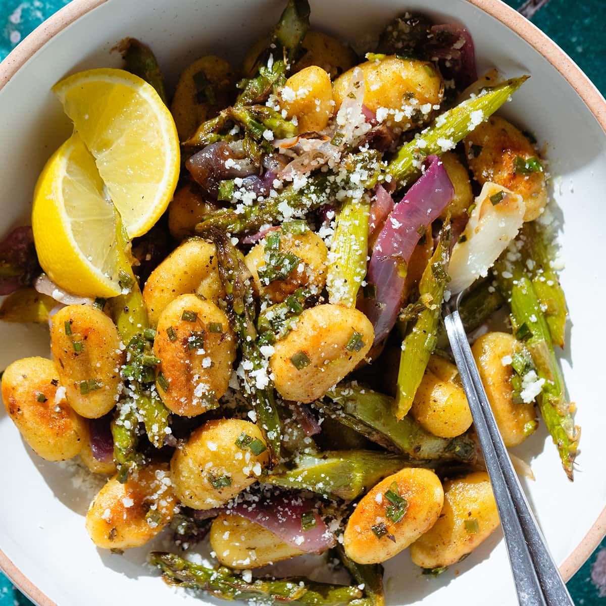 Crispy golden gnocchi with roasted asparagus, red onion, and pecorino in a white bowl with lemon wedges on the side.