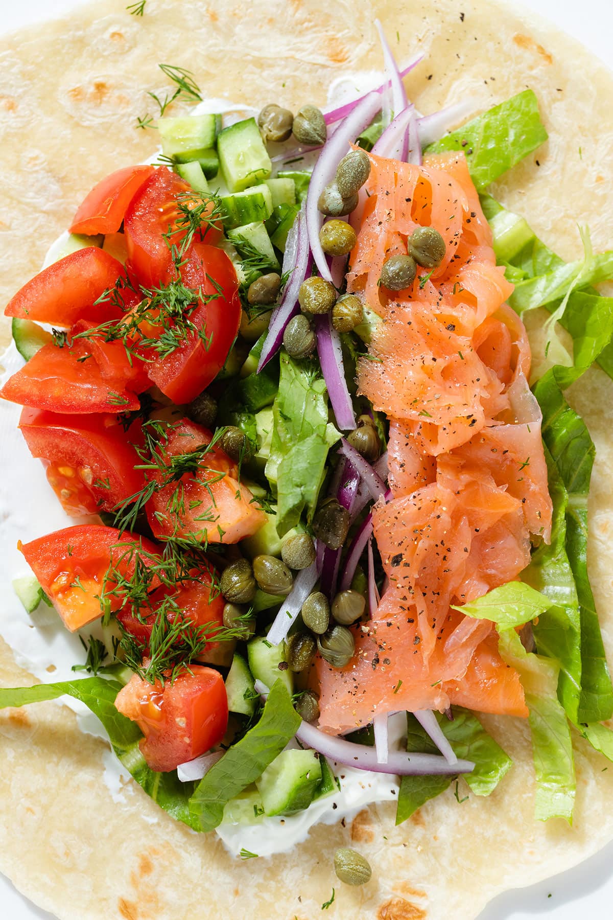 A tortilla wrap topped with chopped tomato, cucumber, smoked salmon, onion, lettuce, and cream cheese.
