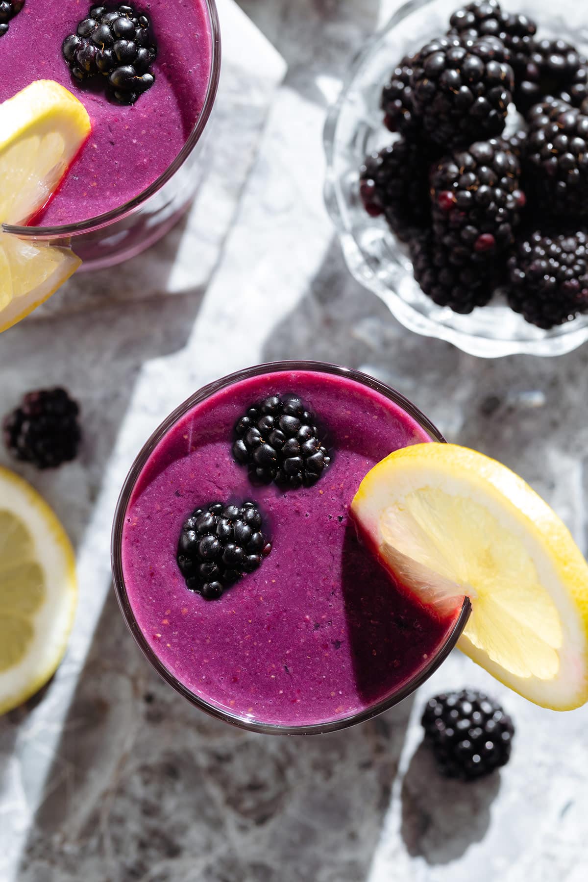 Dark purple smoothie in short glasses garnished with a slice of lemon and fresh blackberries.