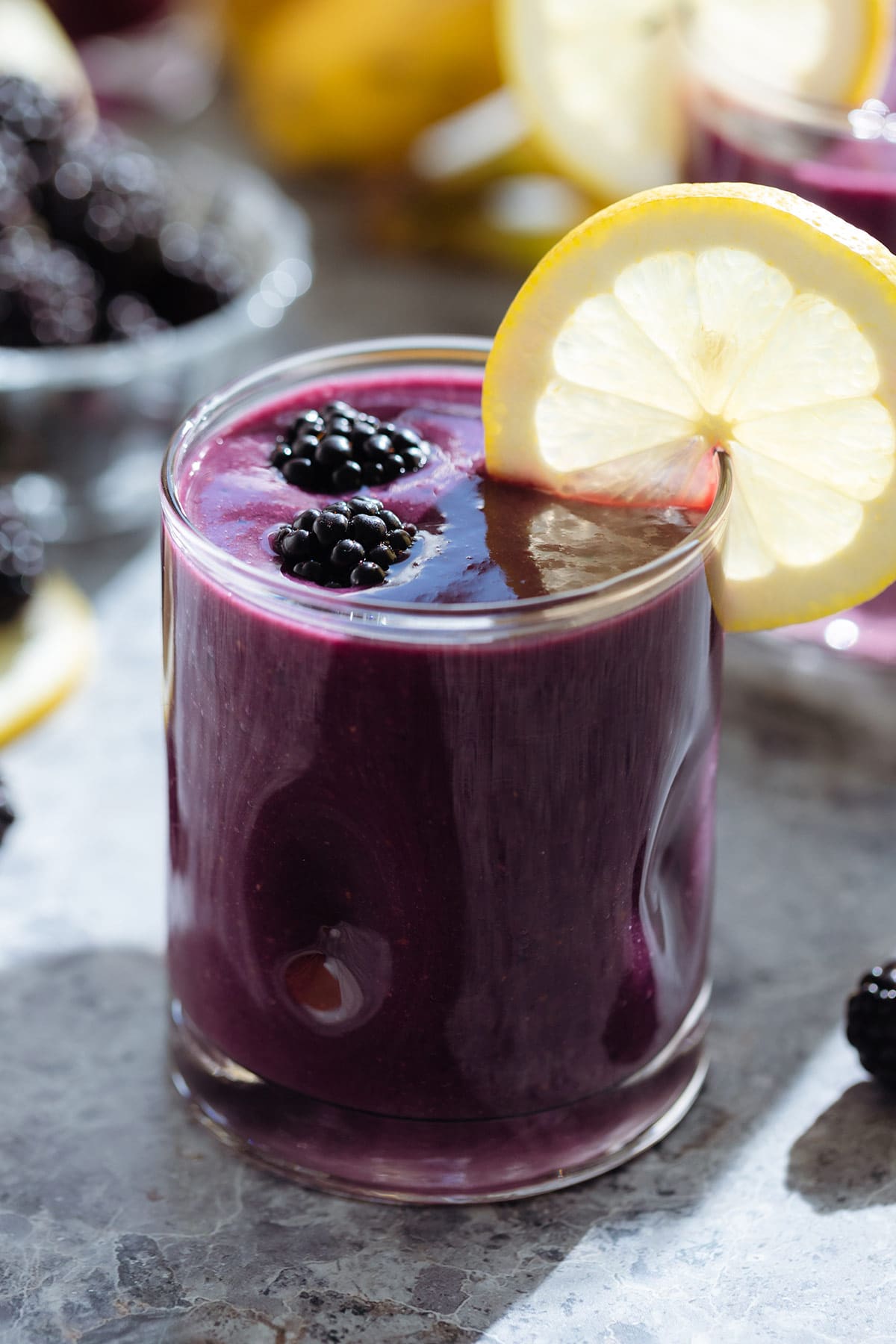 Dark purple smoothie in a short glass garnished with a slice of lemon and fresh blackberries.