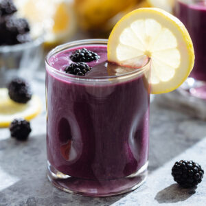 Dark purple smoothie in a short glass garnished with a slice of lemon and fresh blackberries.