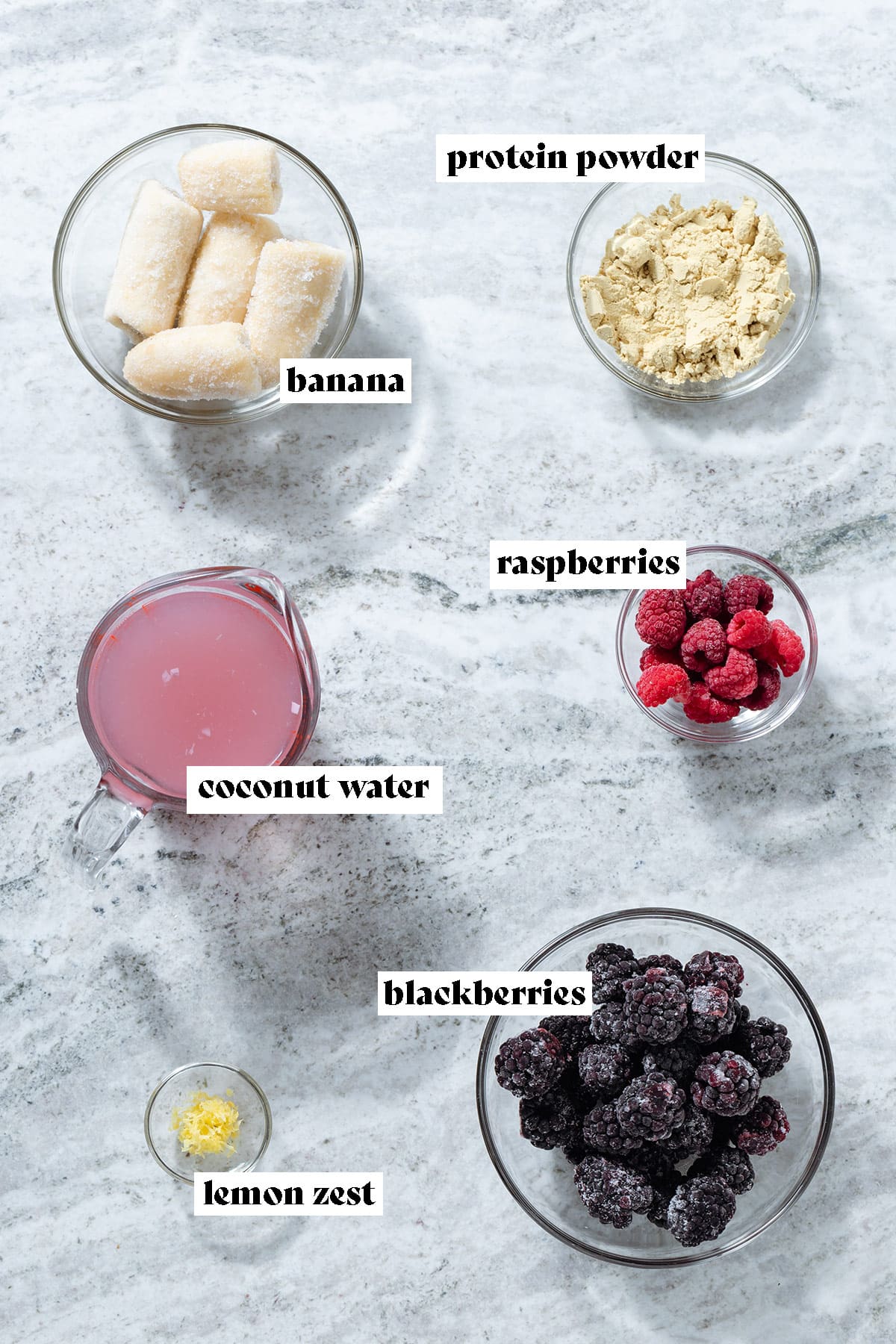 Frozen banana, blackberries, raspberries, coconut water, protein powder, and lemon zest laid out in small glass bowls with text overlay.