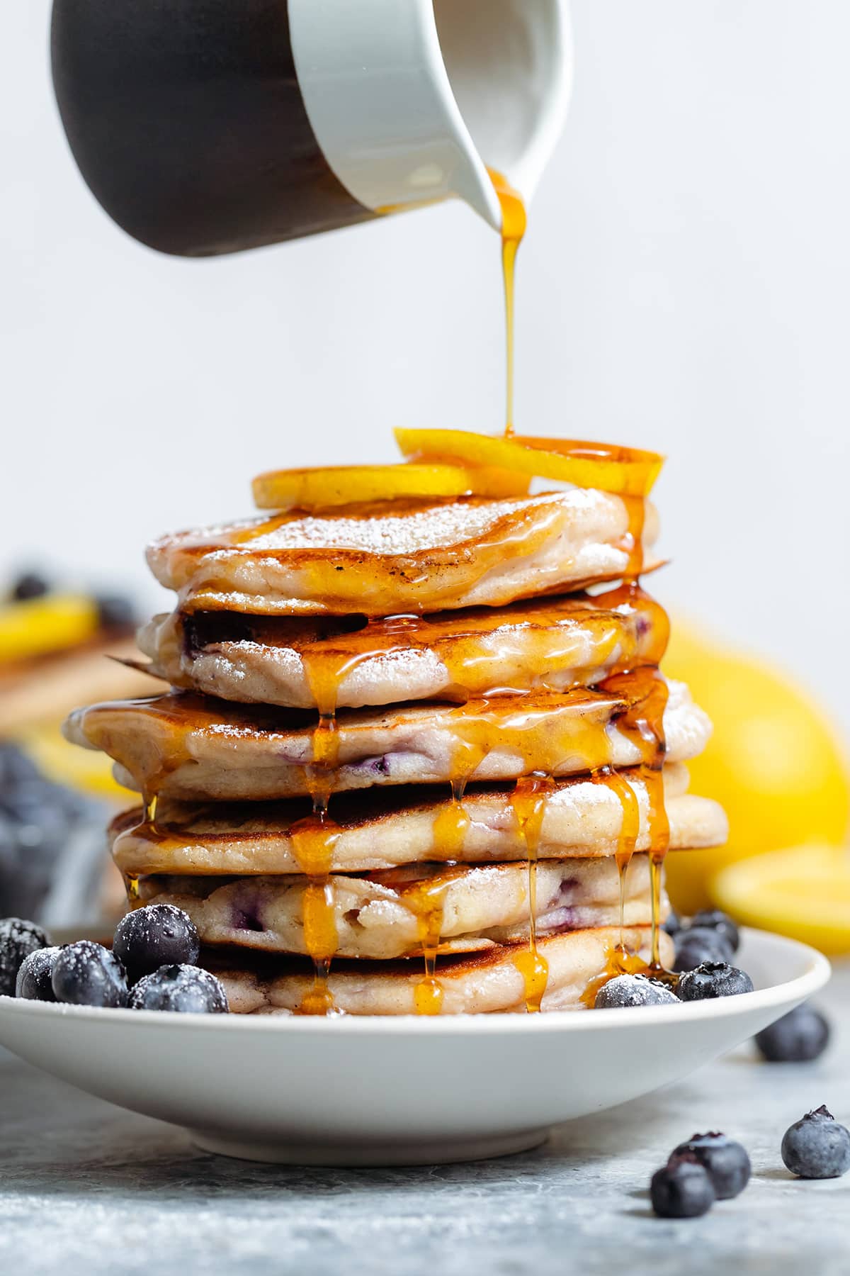 A stack of pancakes garnished with blueberries and lemon slices being drizzled with maple syrup.