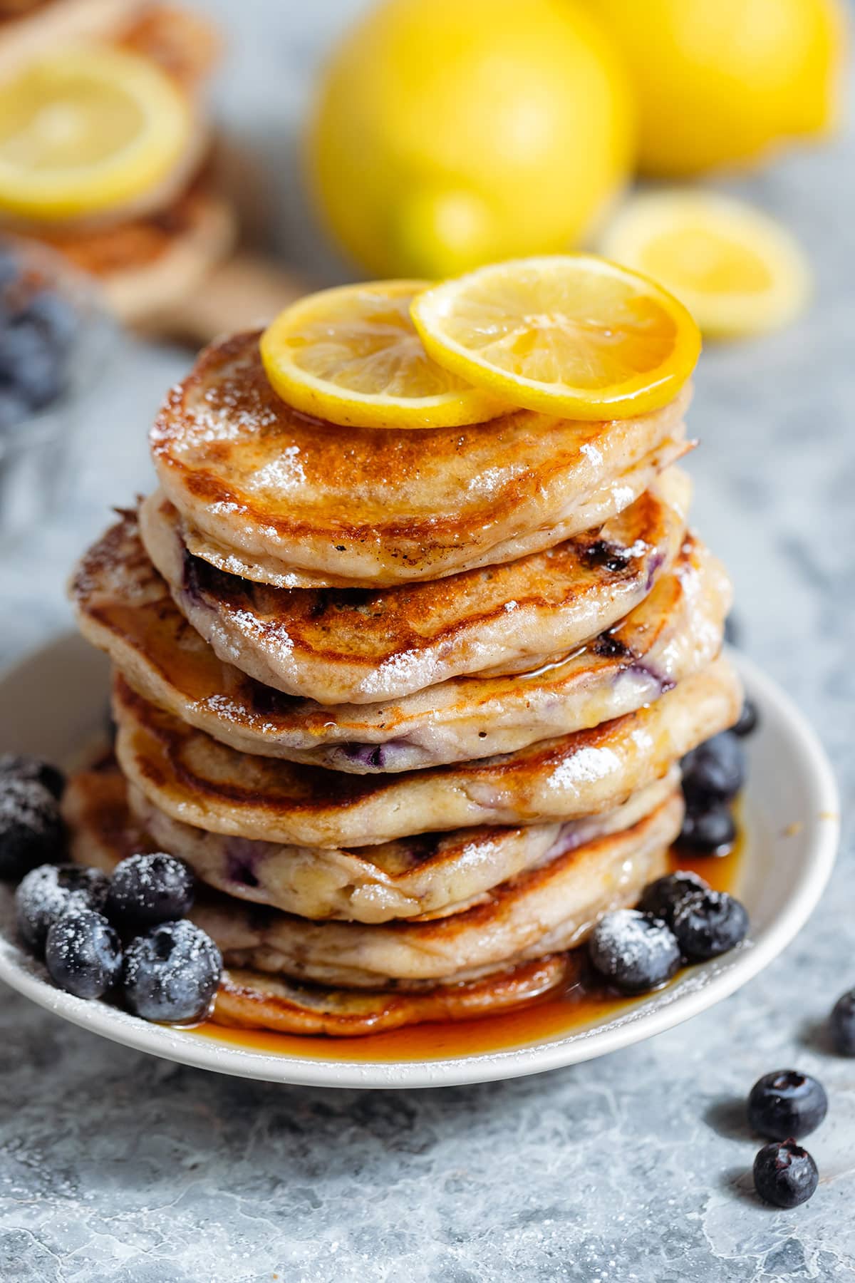 A stack of blueberry pancakes garnished with lemon slices and more blueberries, and drizzled with maple syrup.