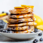 A stack of blueberry pancakes garnished with fresh blueberries, lemon slices, and maple syrup.
