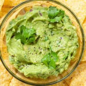 Guacamole in a small glass bowl garnished with fresh cilantro with lots of tortilla chips around the bowl.