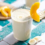 Light yellow smoothie in a tall glass garnished with a slice of banana and peach.