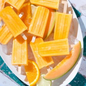 Bright orange cantaloupe popsicles laid out on a large with plate with ice and slices of cantaloupe.