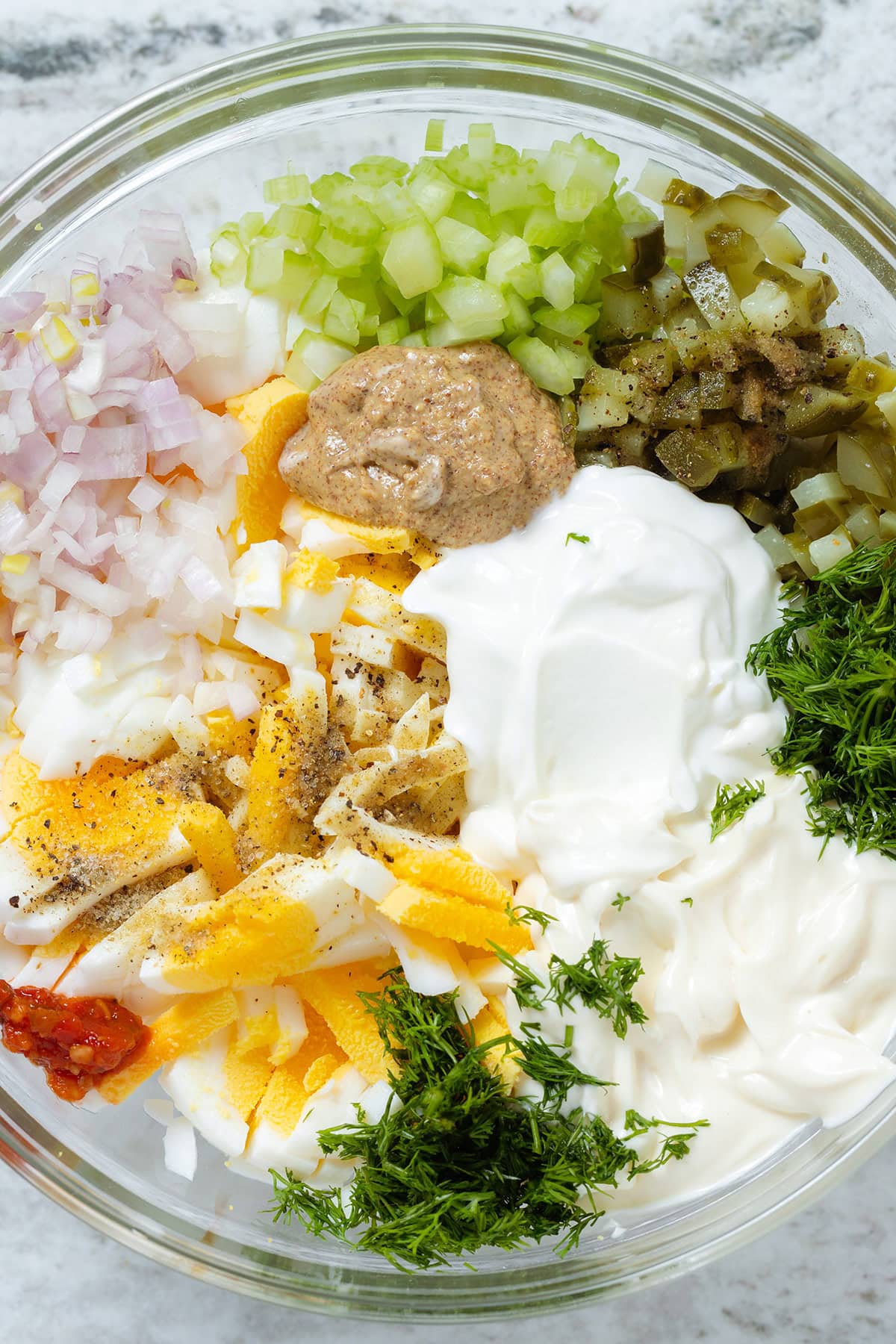 Hardboiled eggs, yogurt, mayonnaise, pickles, dill, celery, and other ingredients in a large glass bowl.