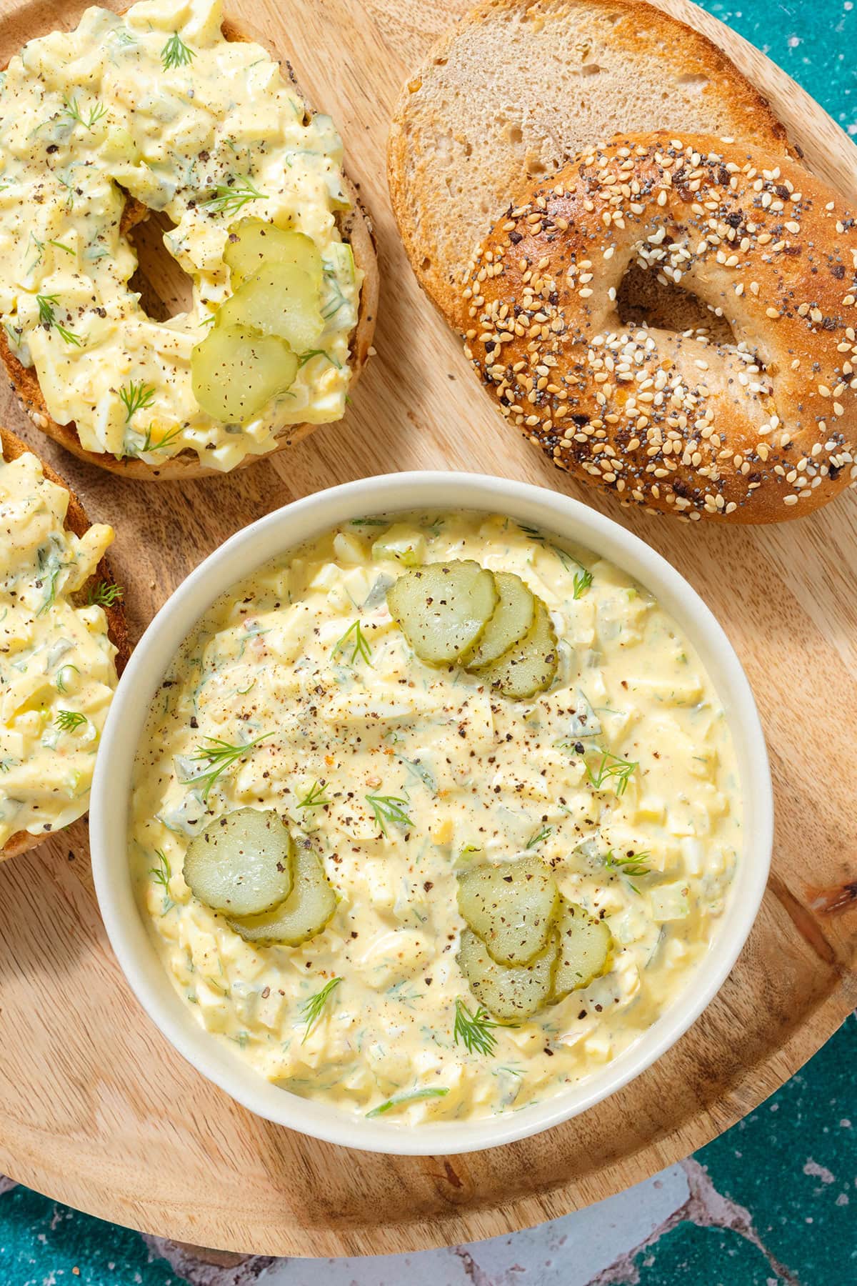 Egg salad in a white bowl garnished with pickles and dill with bagels topped with the egg salad next to it all on a wooden plate.