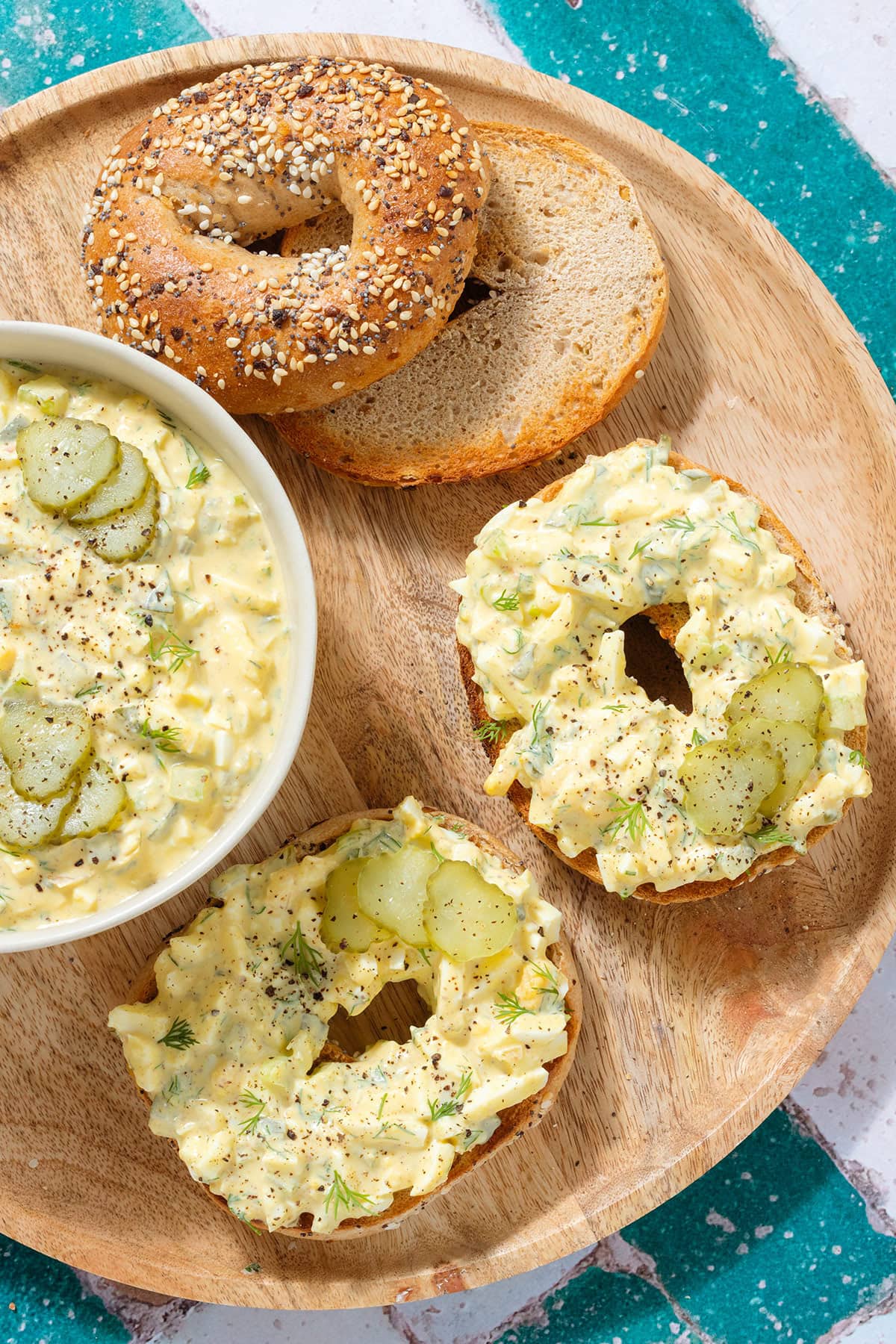 Everything bagels with egg salad and pickle slices on a large wooden plate.