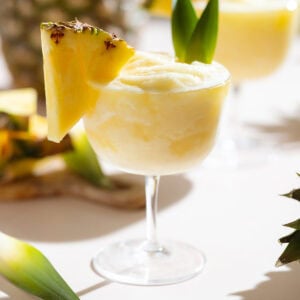Frozen margarita in a coupe glass garnished with fresh pineapple and pineapple leaves.