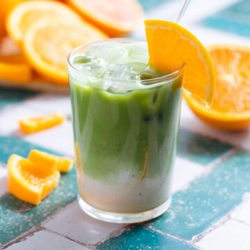 Iced matcha latte in a tall glass garnished with a slice of orange in the glass and on the rim with a glass straw.