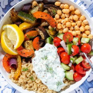 Quinoa bowl with roasted vegetables, chickpeas, fresh salad, and tzatziki in a white bowl.