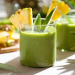 Bright green smoothie in short glasses garnished with slices of pineapple and pineapple leaves.
