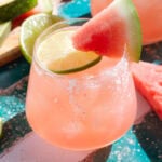 Light red margarita in a short glass with a salt rim garnished with a slice of lime and a wedge of watermelon.
