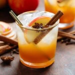 Dark orange iced drink in a short glass with a gold rim with sparkling water on top creating a layered effect, garnished with a cinnamon stick and a star anise.