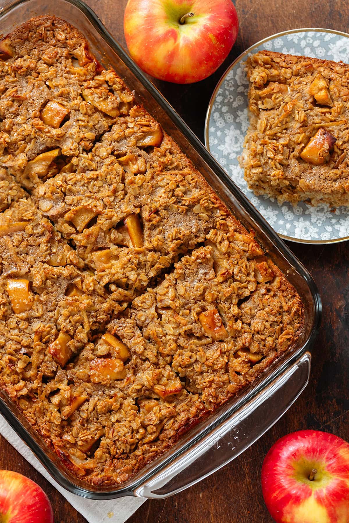 Golden brown baked oatmeal with apples in a large baking dish with one square on a blue plate on the right side of it.