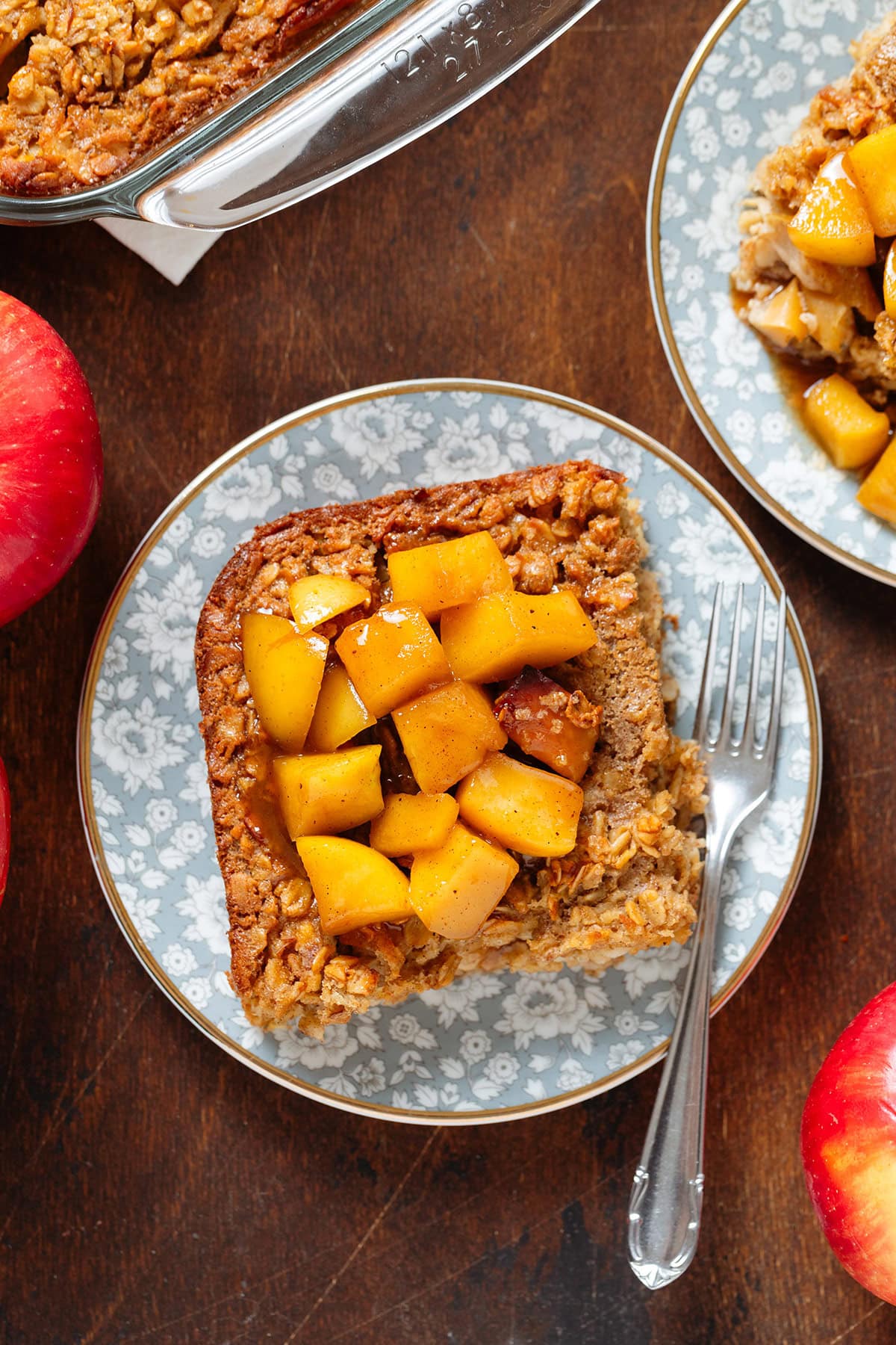 A square slice of apple baked oatmeal on a small blue plate with a gold rim and a small dessert fork on the right side topped with stewed apples.