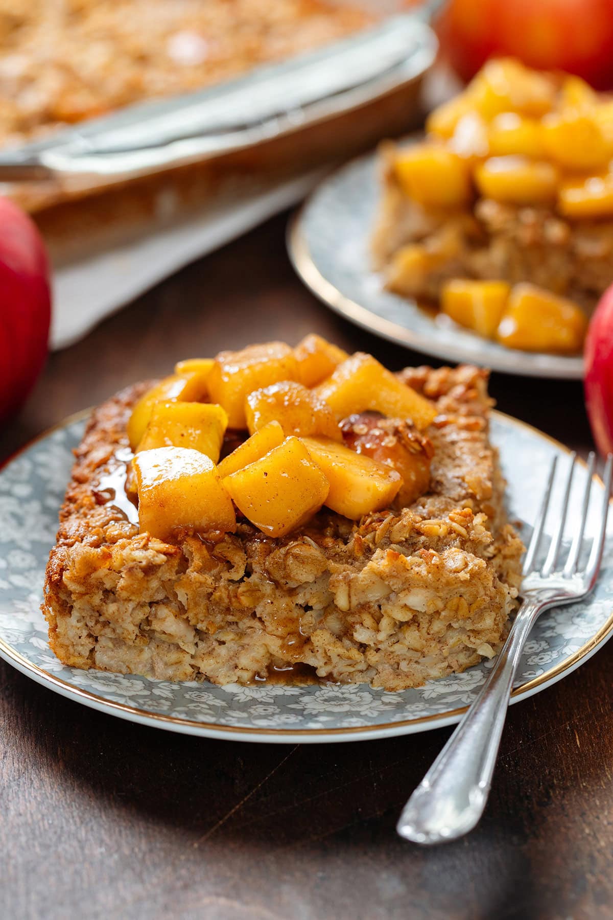 A square slice of apple baked oatmeal on a small blue plate with a gold rim and a small dessert fork on the right side topped with stewed apples and more baked oats in the background.