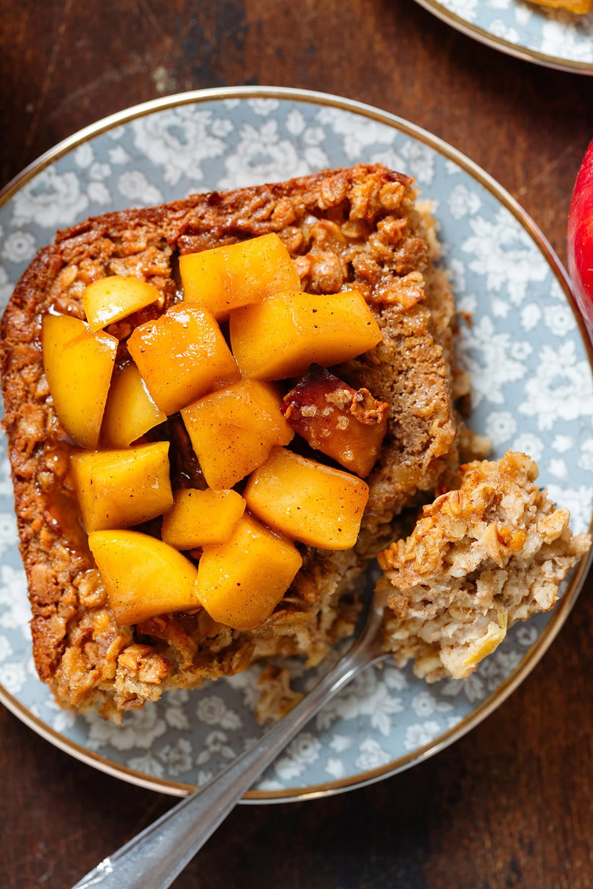 A square slice of apple baked oatmeal on a small blue plate with a gold rim topped with stewed apples and with a small fork holding a bite of the baked oats showing the inside.