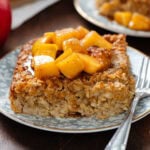 A square slice of apple baked oatmeal on a small blue plate with a gold rim and a small dessert fork on the right side topped with stewed apples.