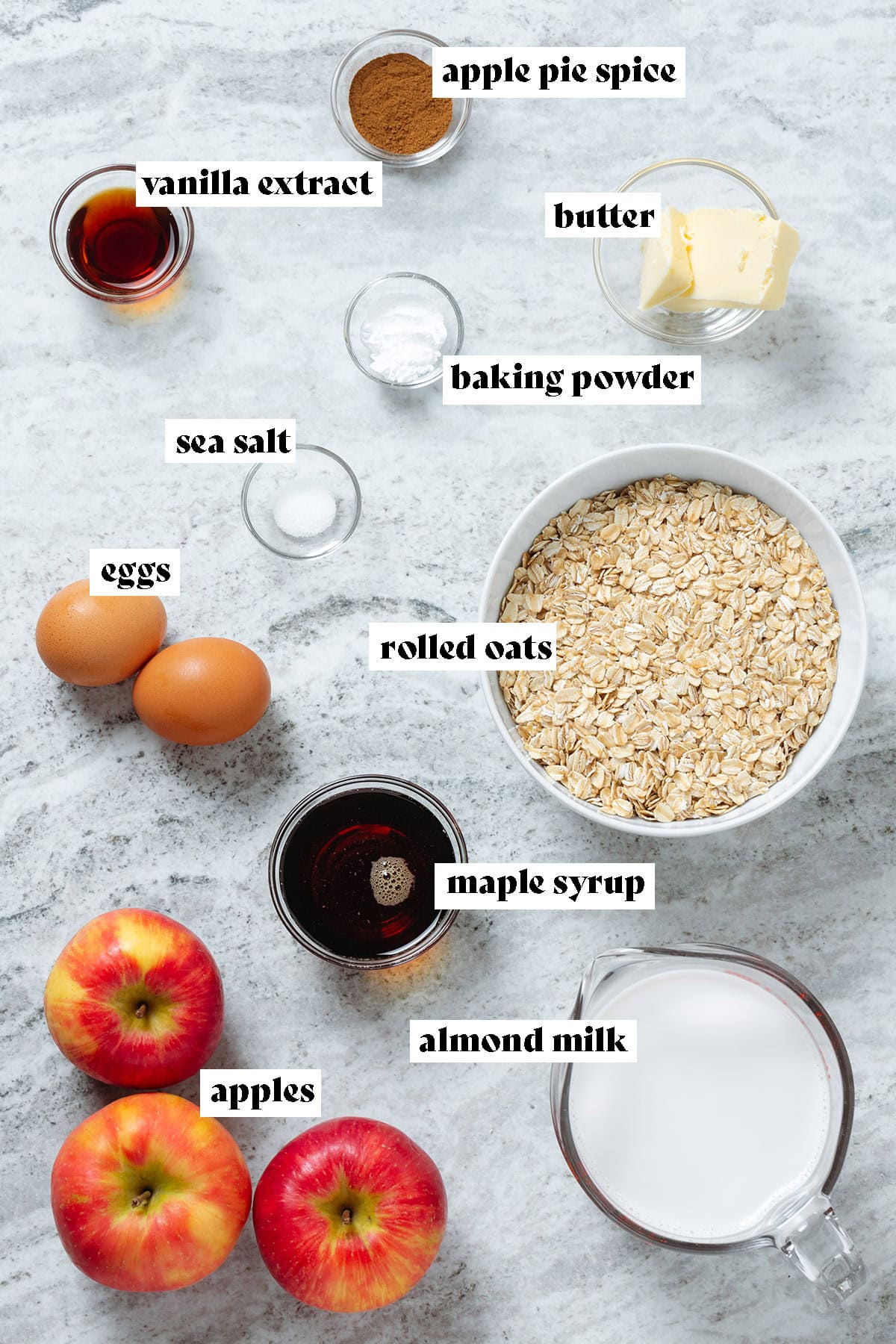 Ingredients like oats, apples, milk, maple syrup, and egg all laid out and some measured in small glass bowls.