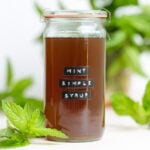 Light brown mint syrup in a glass jar with a black embossed label on it and fresh mint around the jar.