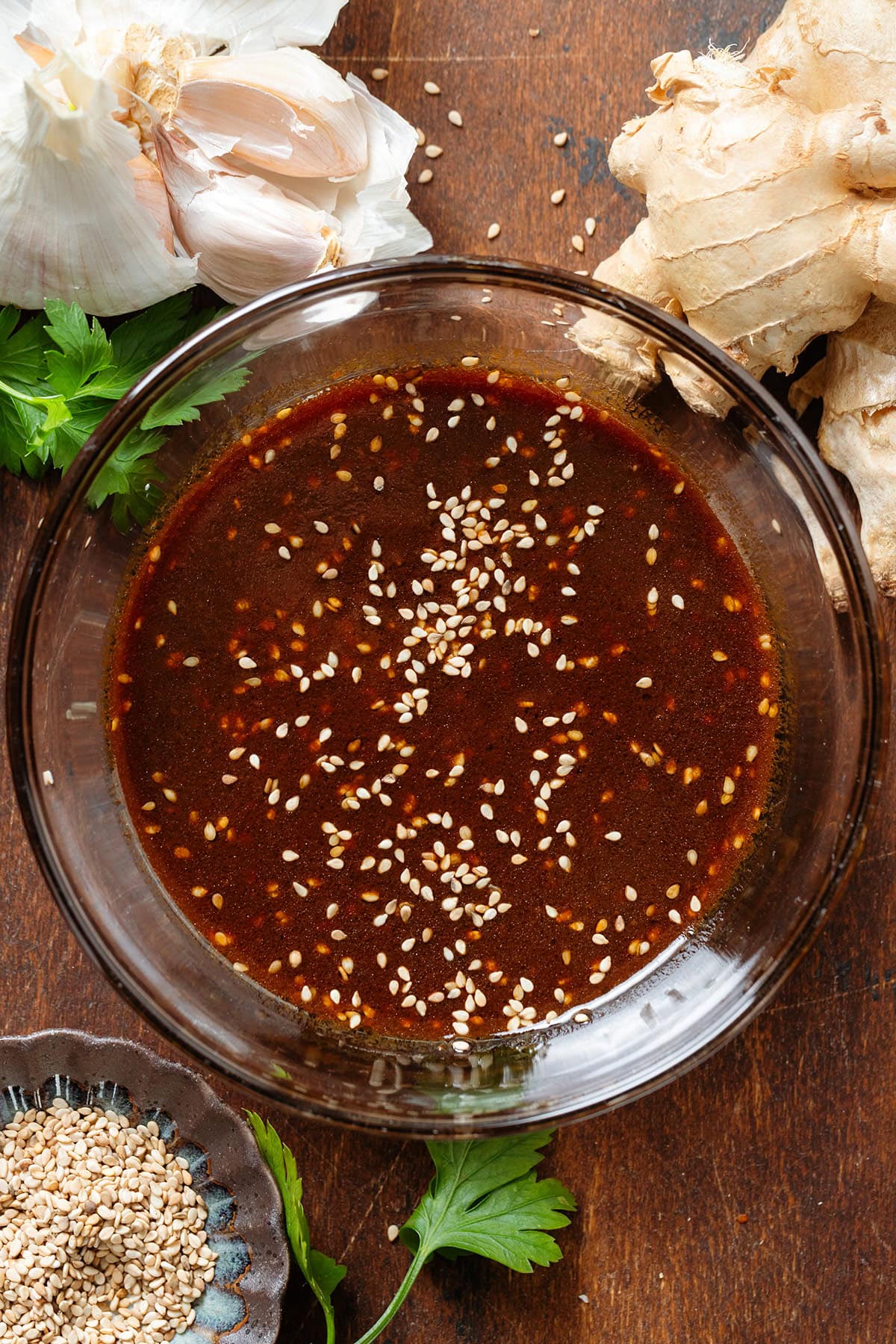 Dark brown miso dressing in a small glass bowl garnished with sesame seeds with fresh ginger, garlic and sesame seeds around the bowl.