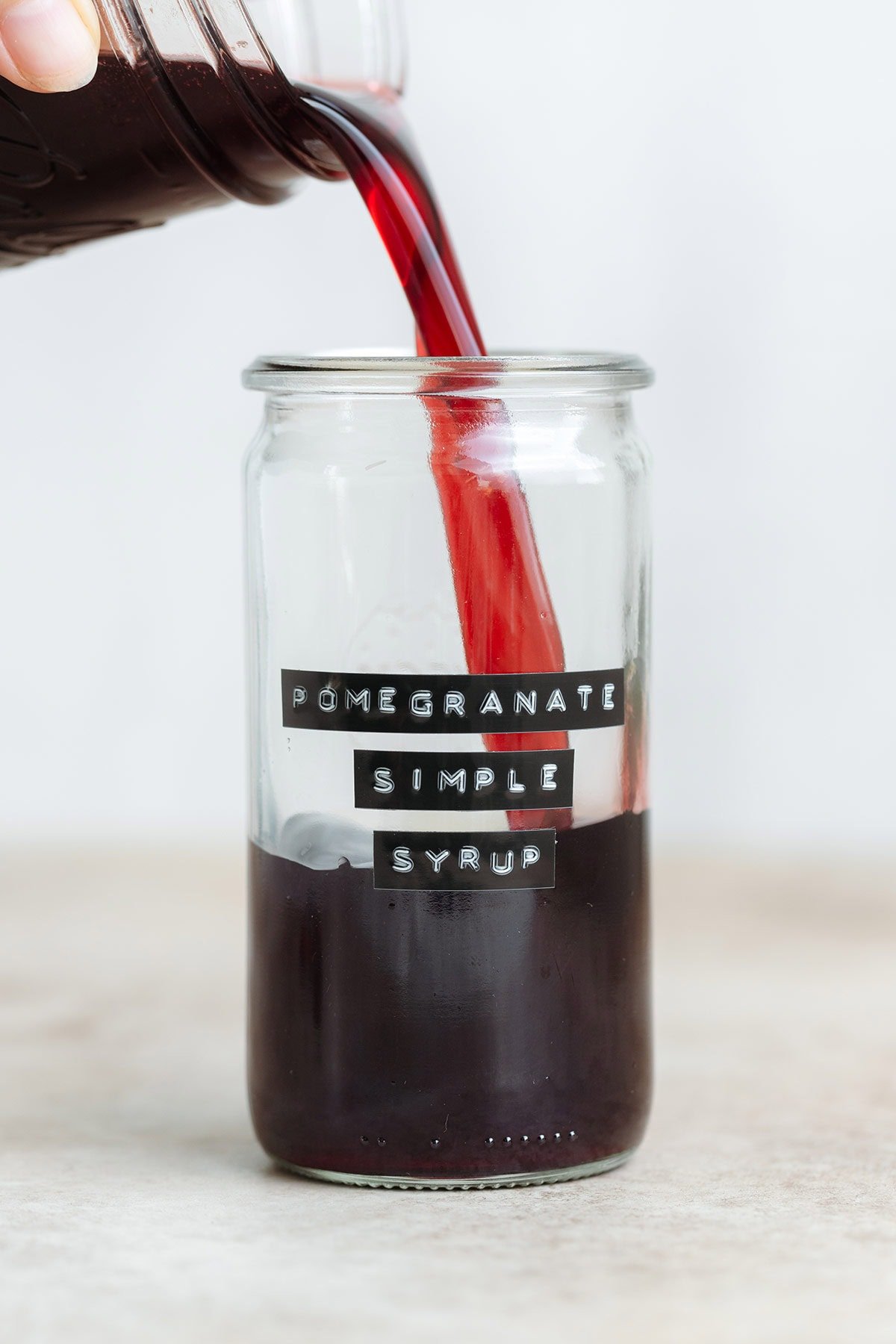 Dark red syrup being poured into a tall glass with an embossed label that says pomegranate simple syrup.