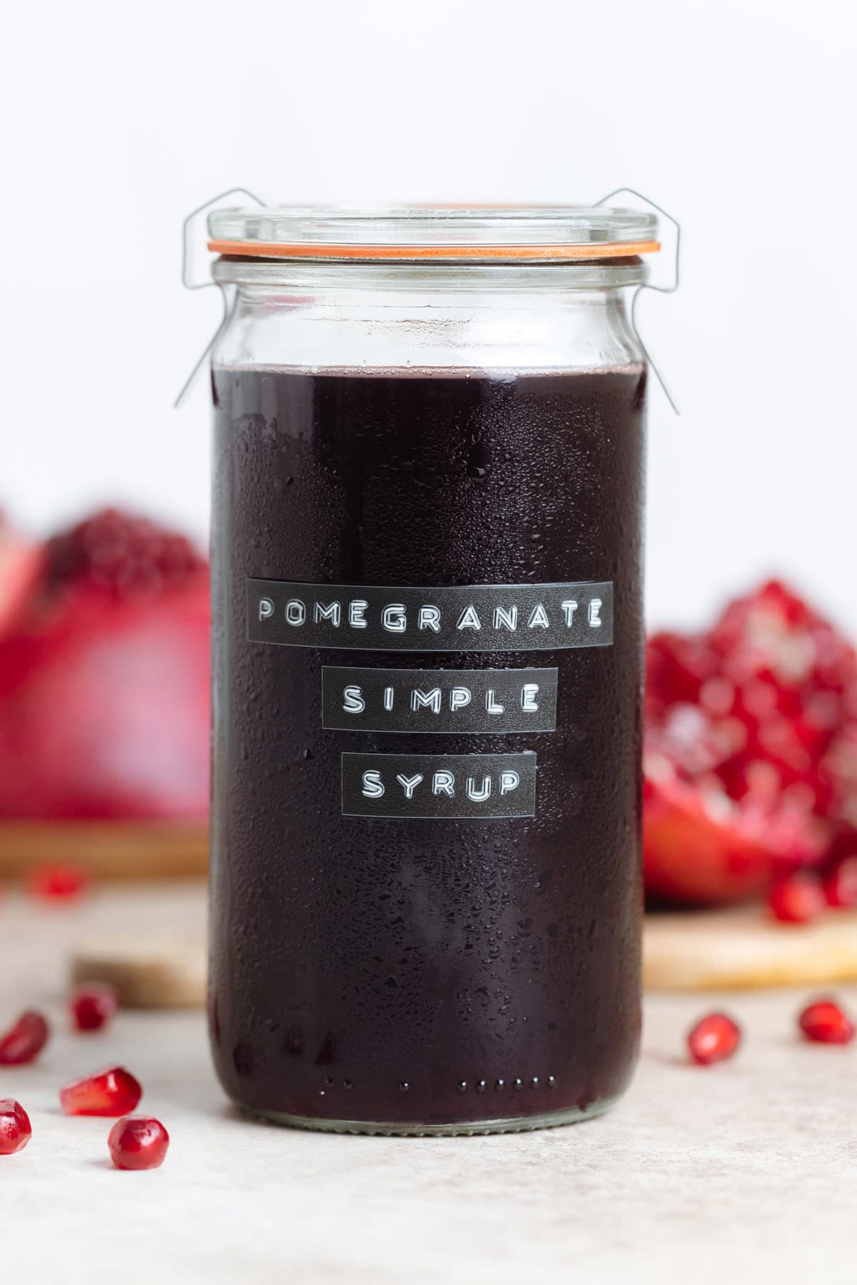 Dark red pomegranate syrup in a tall glass jar with a glass lid with fresh pomegranates around the jar.