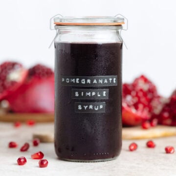 Dark red pomegranate syrup in a tall glass jar with a glass lid with fresh pomegranates around the jar.