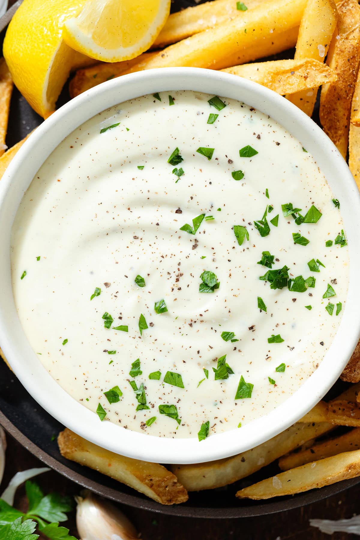 Creamy white dip in a small white bowl garnished with fresh parsley on a black plate with french fries.