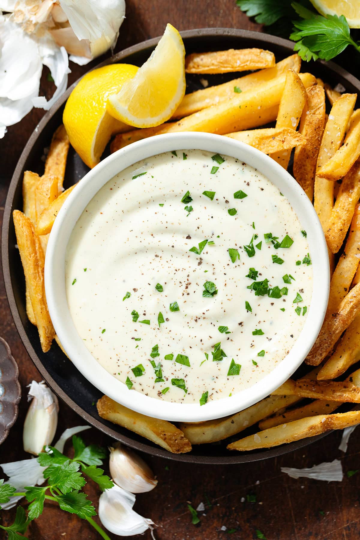 Creamy white dip in a small white bowl garnished with fresh parsley on a black plate with french fries.